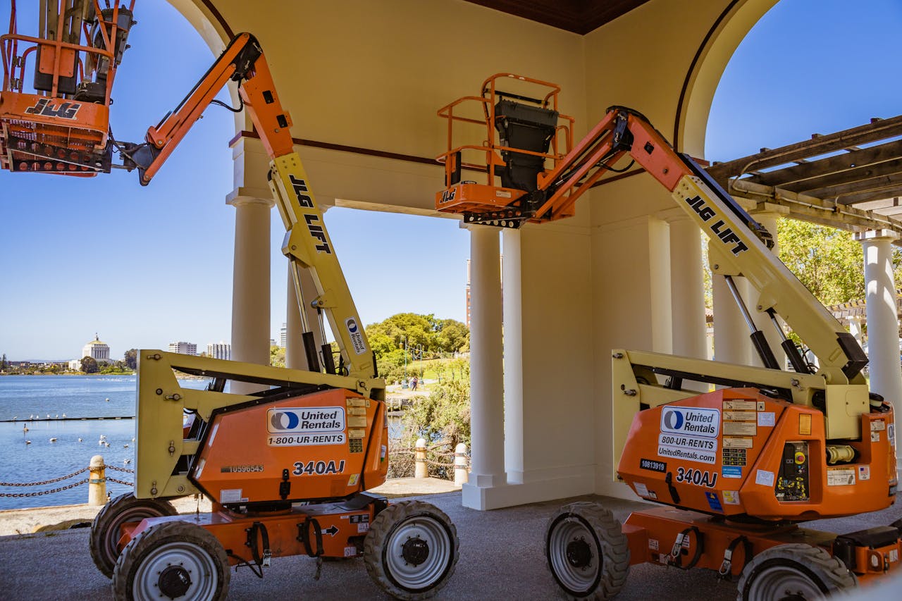 our-journey-03 Orange construction lifts positioned near a lakeside with clear sky and architecture.